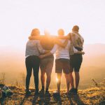 Community Impact - four person hands wrap around shoulders while looking at sunset